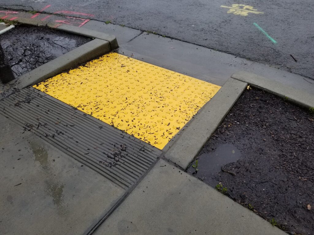 A picture of a brightly painted curb cut with tactile dots against plain concrete.